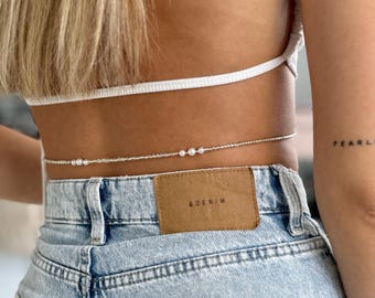 May include: A thin waist chain featuring small silver-toned beads and three clusters of white pearls, worn over light blue denim jeans with a tan leather patch labeled & DENIM and a white ribbed top with a horizontal strap across the back.
