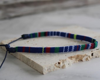 Peut inclure: Un collier ras du cou bleu tissé avec un motif rayé rouge, vert et blanc. Le collier a un cordon bleu foncé avec une fermeture à nœud. Le choker est présenté sur une surface claire et texturée.