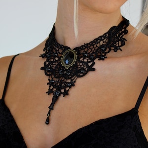 Puede incluir: Collar gargantilla de encaje negro con un colgante ovalado de piedra oscura. El intrincado diseño presenta detalles de cuentas y un colgante en forma de lágrima. El collar se lleva sobre un top negro.