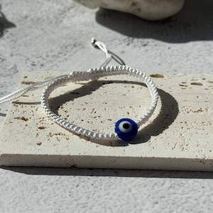 May include: White macrame bracelet featuring a blue evil eye bead. The bracelet is adjustable with sliding knots and is displayed on a textured, light-colored surface. The evil eye bead is a deep blue with a white and black center.