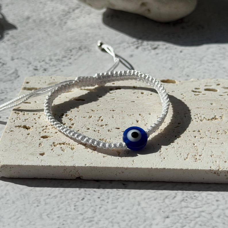 May include: White macrame bracelet featuring a blue evil eye bead. The bracelet is adjustable with sliding knots and is displayed on a textured, light-colored surface. The evil eye bead is a deep blue with a white and black center.
