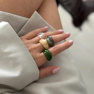 May include: Five chunky resin rings in clear, beige, marbled gray, and green hues. The rings are worn on the fingers, showcasing their varied colors and glossy finish. The rings are a fashion accessory.
