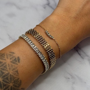 Op de afbeelding: Een collectie gouden armbanden siert een pols. De armbanden omvatten een armband met baguette-geslepen diamanten, een geweven gouden armband en een delicate kettingarmband met kleine edelstenen. De sieraden worden gepresenteerd op een lichte huidskleur.