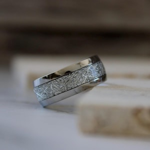 May include: A silver-colored ring with a textured, patterned center band. The ring has a polished, metallic finish and a modern design. The ring is likely made of a durable metal, such as stainless steel or titanium.