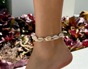 May include: A double-strand anklet featuring a row of natural white cowrie shells and one metallic gold-toned cowrie shell. The shells are strung on a thin, light-colored cord that wraps around the ankle, creating a layered appearance.