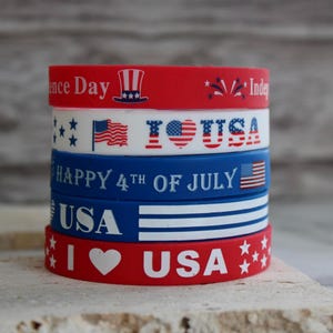 May include: A stack of five patriotic rubber wristbands in red, white, and blue. The bands feature text and graphics including the American flag, stars, and the words "USA" and "Happy 4th of July".