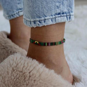 May include: A colorful woven ankle bracelet with a geometric pattern in green, red, yellow, and purple. The bracelet is worn on an ankle, with blue jeans and a fluffy slipper in the background.