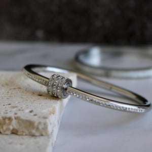 May include: A silver-toned bangle bracelet with a central cylindrical design adorned with multiple rows of small, clear crystals. The bracelet's band is also embellished with crystals. Another bracelet is visible in the background.
