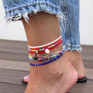 Può includere: Braccialetti alla caviglia in vari colori. I braccialetti sono fatti di piccole perline colorate e presentano un nappa e una conchiglia bianca. I braccialetti sono indossati sulla caviglia di una persona, con jeans di denim sullo sfondo.