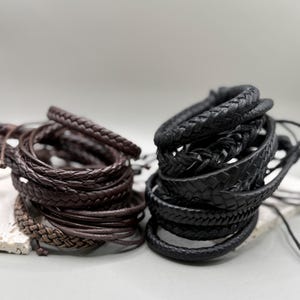 Pode incluir: Uma coleção de pulseiras de couro trançado dispostas em duas pilhas distintas. Uma pilha consiste em vários cordões e fios trançados de couro marrom escuro, enquanto a outra pilha apresenta vários cordões e faixas trançadas de couro preto.