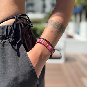 Puede incluir: Una persona lleva una pulsera tejida de color negro y rosa con una pulsera de cuero marrón en la muñeca. La pulsera tiene un pequeño dije de oro.