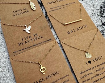 Puede incluir: Conjunto de cinco collares de oro con diferentes dijes y mensajes inspiradores en cartulina marrón. Los dijes incluyen una pluma, un pájaro, una clave de sol, una barra y un corazón. Los mensajes incluyen "Light as a Feather", "Life is Beautiful", "Make Music", "Balance" y "I Love You!"