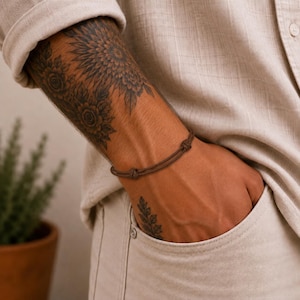 Può includere: Un semplice bracciale in corda marrone con due nodi scorrevoli regolabili. Il bracciale è composto da due sottili fili paralleli di materiale marrone scuro che avvolgono il polso, creando un aspetto pulito e sobrio per l'uso quotidiano.