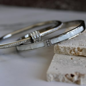 Puede incluir: Tres pulseras plateadas. Una está incrustada con pequeños cristales, otra tiene una incrustación rectangular blanca con tres pequeños cristales, y la tercera es una simple banda plateada. Las pulseras descansan sobre una superficie clara y texturizada.