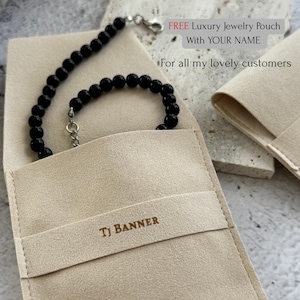 May include: A black beaded bracelet with a silver clasp rests inside a beige jewelry pouch. The pouch is embossed with the text "TJ BANNER". The image also includes text that reads "FREE Luxury Jewelry Pouch With YOUR NAME".