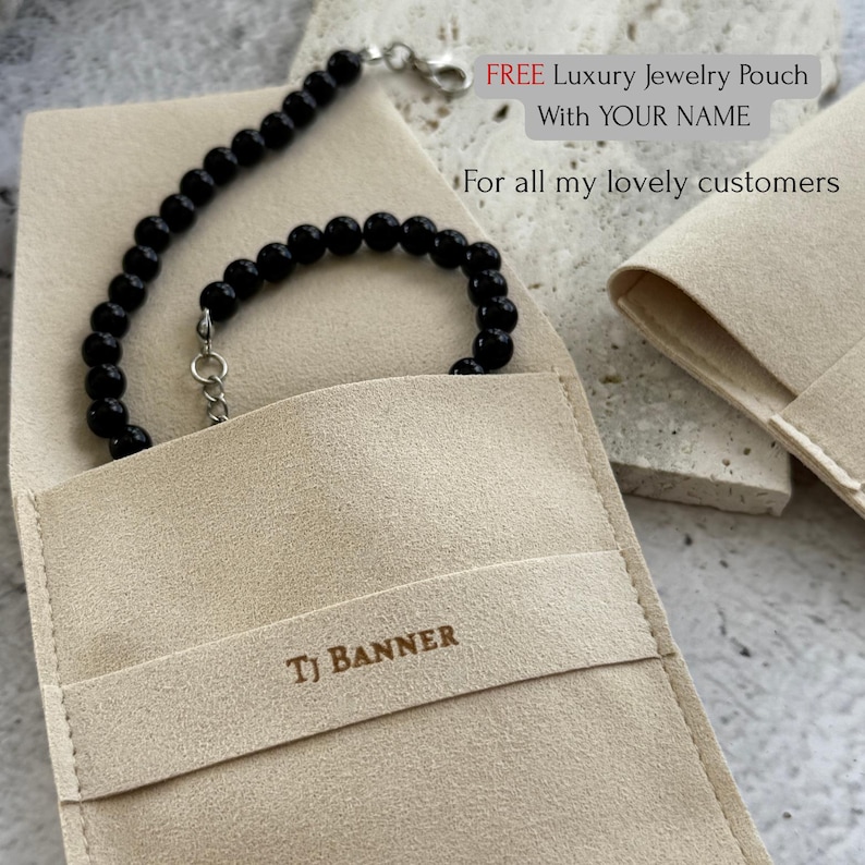 May include: A black beaded bracelet with a silver clasp rests inside a beige jewelry pouch. The pouch is embossed with the text "TJ BANNER". The image also includes text that reads "FREE Luxury Jewelry Pouch With YOUR NAME".