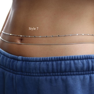 May include: A double-strand silver-toned waist chain featuring one delicate textured chain and one smooth snake chain. The text "Style 7" is visible above the chains. The jewelry is worn around the midsection above blue sweatpants.