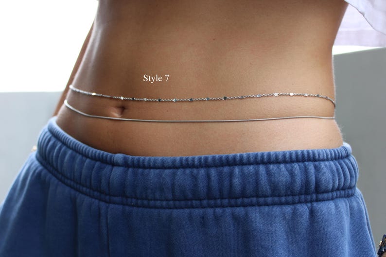 May include: A double-strand silver-toned waist chain featuring one delicate textured chain and one smooth snake chain. The text "Style 7" is visible above the chains. The jewelry is worn around the midsection above blue sweatpants.