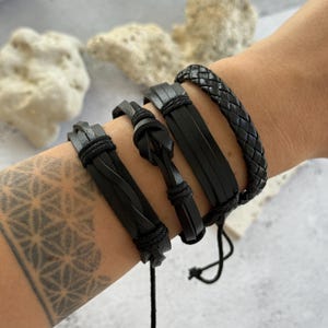 May include: A set of four black leather bracelets worn together. The collection includes a braided leather band, a wide flat leather strap, a corded band with a central knot detail, and a thin leather strip with a twisted design.