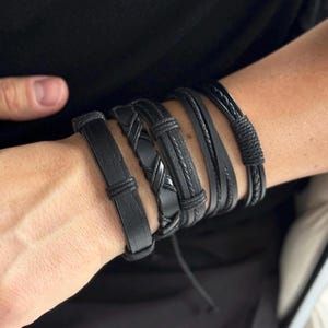 Może przedstawiać: Zestaw pięciu czarnych skórzanych bransoletek z różnymi wzorami plecionymi i tkanymi. Bransoletki są ułożone na nadgarstku osoby.