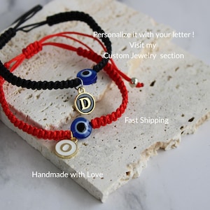 May include: Two handmade bracelets, one black and one red, with an evil eye charm and a letter pendant. The bracelets are on a textured surface. The image includes the text "Personalize it with your letter!" and "Handmade with Love."