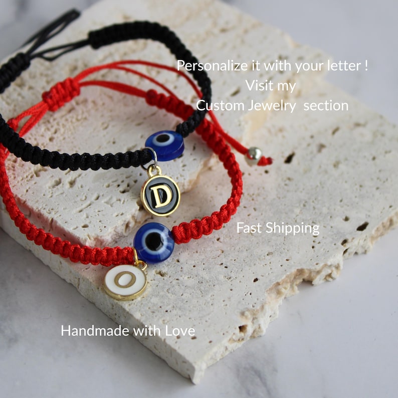 May include: Two handmade bracelets, one black and one red, with an evil eye charm and a letter pendant. The bracelets are on a textured surface. The image includes the text "Personalize it with your letter!" and "Handmade with Love."