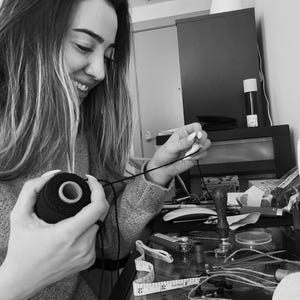 May include: A black and white image of a person working with black thread and a spool. The person is holding the spool of thread in one hand and manipulating the thread with the other. A measuring tape and other crafting tools are on the table.