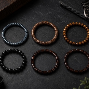 May include: Six circular bracelets arranged in two rows. The top row features a grey disc-bead bracelet, a textured brown wood-grain bracelet, and a smooth brown bead bracelet. The bottom row includes a matte black bead bracelet and two dark brown cylindrical bead bracelets.