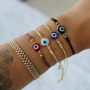 May include: A collection of bracelets featuring gold and silver metal watch-style bands and string bracelets with evil eye charms in red, blue, and black. The bracelets are displayed on a person's wrist.