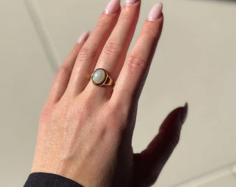 May include: A gold ring with a white oval gemstone. The ring is on a person's finger.