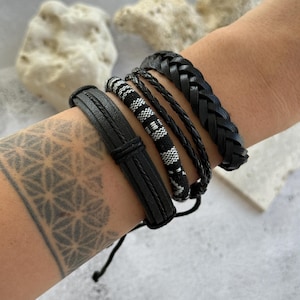 May include: A selection of black bracelets in different designs. The bracelets include a plaited leather band, a woven band with black and white patterns, and several thin, braided bands. The bracelets are shown on a person's arm.