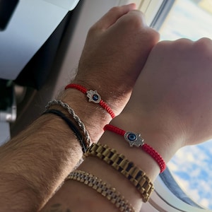 May include: Two red string bracelets with silver Hamsa hand charms, each featuring a blue eye. Other bracelets include a silver rope chain and a gold watch. The bracelets are worn on wrists.