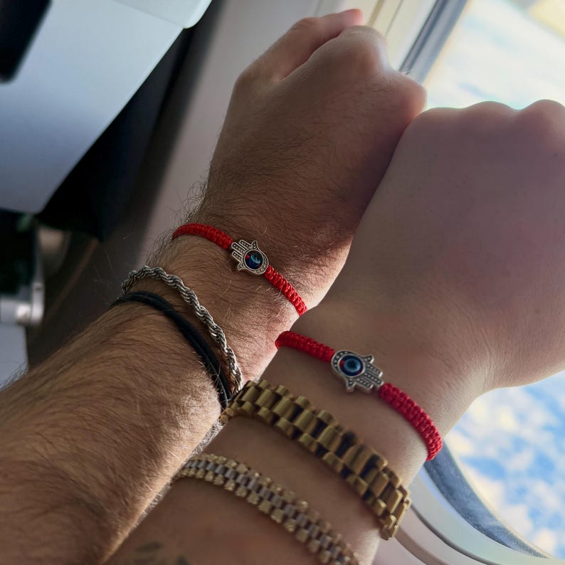 May include: Two red string bracelets with silver Hamsa hand charms, each featuring a blue eye. Other bracelets include a silver rope chain and a gold watch. The bracelets are worn on wrists.