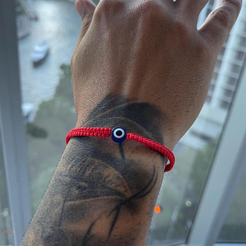 May include: A red woven bracelet with a blue and white evil eye charm. The bracelet is tied around the wrist. The charm is a small, round bead with a blue center, white iris, and black pupil.
