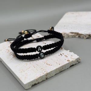 Pode incluir: Duas pulseiras de amizade pretas com as letras "A" e "S" nelas. As pulseiras são feitas de cordão preto e estão amarradas juntas.