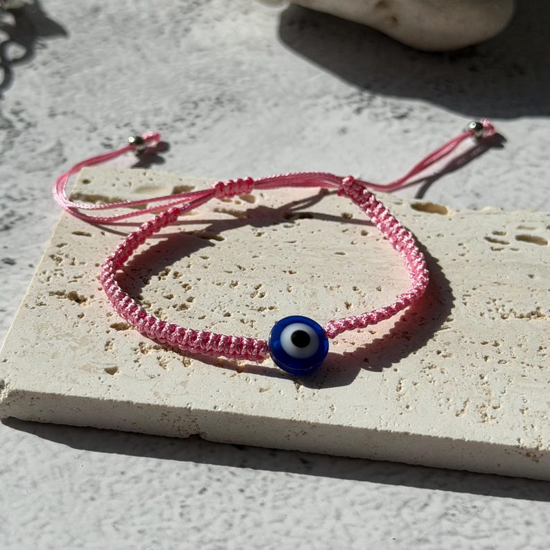 May include: Pink woven bracelet featuring a blue and white evil eye charm. The bracelet is made of pink cord and has an adjustable sliding knot closure. The evil eye charm is a dark blue bead with a white and black eye design.