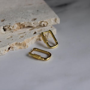 May include: A pair of gold-colored, rectangular hoop earrings. Each earring features a textured, ridged design on the lower portion. The earrings are displayed on a white surface with a textured, light-colored stone in the background.