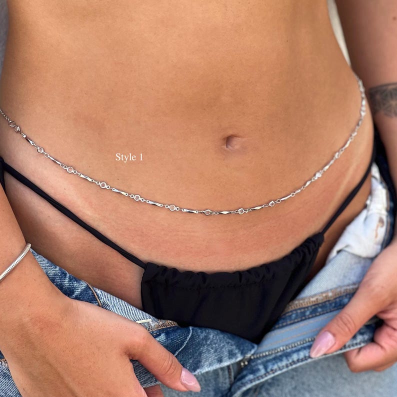 May include: A silver-toned waist chain featuring a delicate link design with small, evenly spaced circular accents. The chain rests across the midsection and includes the text Style 1 printed in white near the center of the jewelry piece.