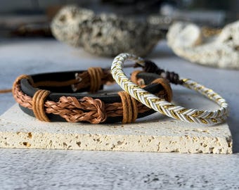May include: Two bracelets on a light-colored surface. One is black leather with brown braided and wrapped accents. The other is white and gold braided. The bracelets are likely for wrist wear.