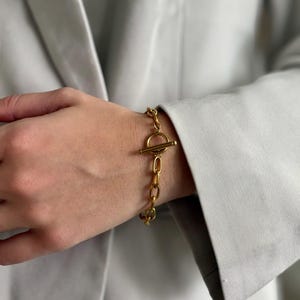 May include: Gold-toned chain bracelet with a toggle clasp. The bracelet features a series of oval links connected by a circular clasp with a horizontal bar. The jewelry is worn on a wrist, with a light gray blazer in the background.