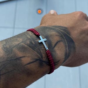 Könnte beinhalten: Ein burgunderfarbenes geflochtenes Armband mit einem silbernen Kreuzanhänger. Das Armband wird an einem Handgelenk mit einem Tattoo getragen. Das Kreuz ist ein schlichtes, elegantes Design, und das Armband besteht aus gewebtem Material.
