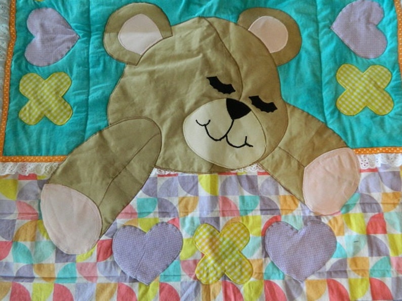 May include: A turquoise and white patchwork quilt with a brown teddy bear applique. The bear has pink paws and a closed-eye expression. The quilt features a variety of shapes, including hearts and crosses.