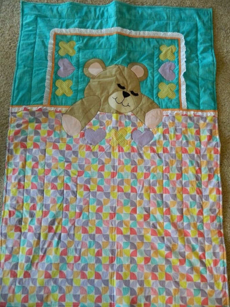 May include: A turquoise and white quilt with a brown teddy bear applique. The quilt has a pattern of colorful half circles.