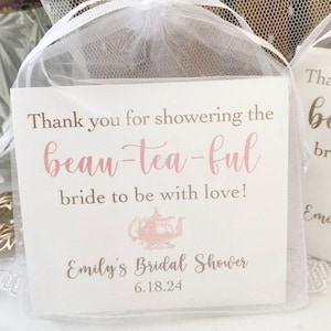 May include: Thank you for showering the beau-tea-ful bride to be with love!  Emily's Bridal Shower 6.18.24.  A white card with pink and brown text in a clear organza bag.