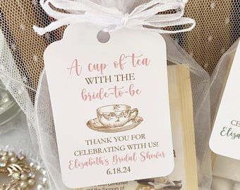 Puede incluir: Una etiqueta de papel rectangular blanca con esquinas redondeadas, atada con una cinta blanca. El texto dice A cup of tea WITH THE bride-to-be, THANK YOU FOR CELEBRATING WITH US! Elizabeth's Bridal Shower 18/06/24, con una ilustración de una taza.