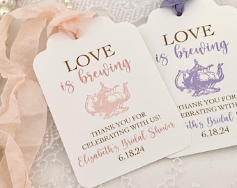 May include: Two white bridal shower favor tags with a pink and purple teapot illustration. The text on the tags reads "Love is brewing" and "Thank you for celebrating with us! Elizabeth's Bridal Shower 6.18.24".