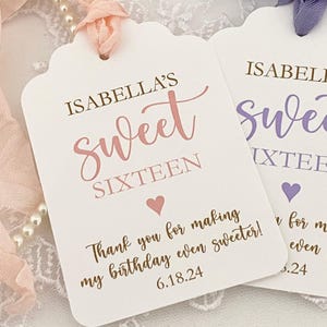 May include: Two white gift tags with scalloped edges. The top tag has pink text that reads "Isabella's Sweet Sixteen" with a small pink heart. The bottom tag has brown text that reads "Thank you for making my birthday even sweeter! 6.18.24". The bottom tag has purple text that reads "Isabella's Sweet Sixteen" with a small purple heart. The bottom tag has brown text that reads "Thank you for making my birthday even sweeter! 6.18.24".