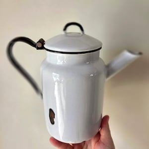 May include: White enamelware teapot with a black handle and spout. The teapot has a lid with a black handle and a small brown spot on the side. The teapot is held by a hand.
