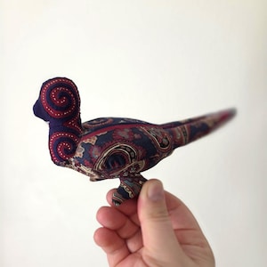 May include: A fabric bird with a paisley pattern in navy blue, burgundy, and cream. The bird has a rounded body, a small head with a spiral design, and a long tail. The bird is held in a hand against a white background.