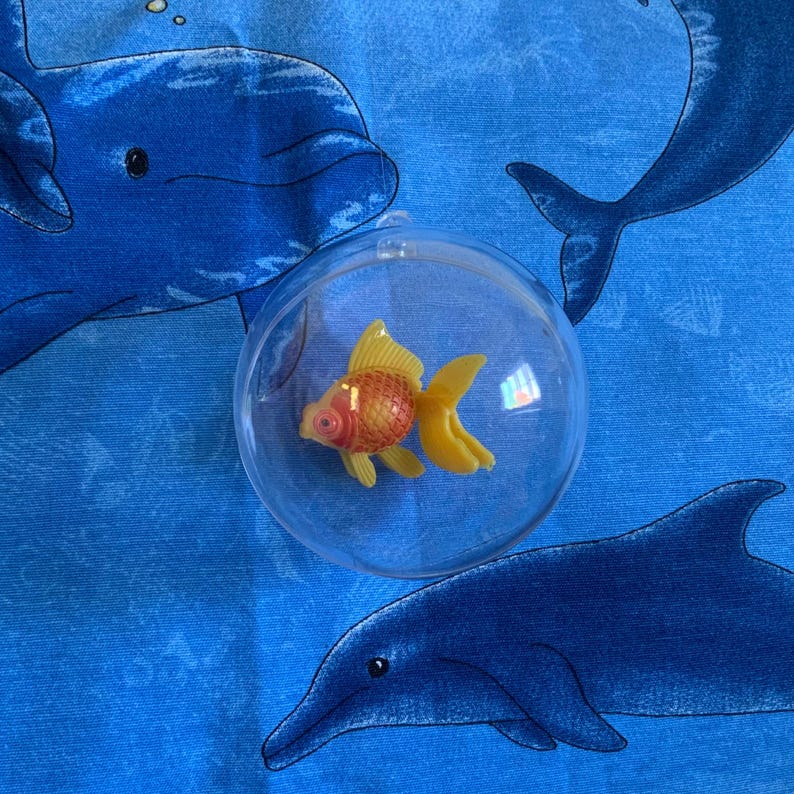 Puede incluir: Un adorno de pl&aacute;stico transparente que contiene un peque&ntilde;o pez dorado naranja y amarillo. El adorno est&aacute; sobre un fondo azul con patrones de delfines.
